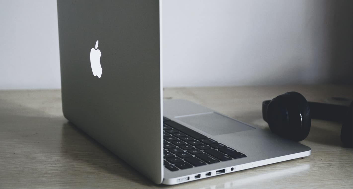 Silver laptop with an illuminated apple logo on a wooden desk, next to black headphones.