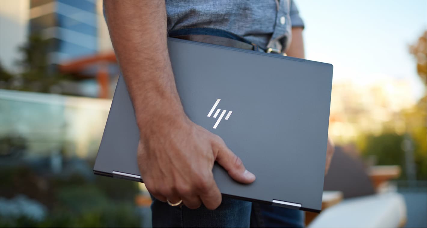 Person holding a closed HP laptop outdoors, wearing a casual shirt and jeans, with a blurred urban background.