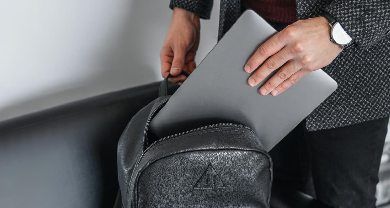 Person placing a silver laptop into a black leather backpack, with a focus on their hands and watch.