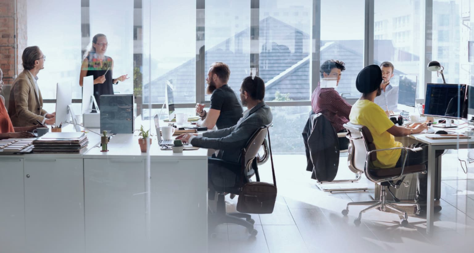 Open-plan office with people working at desks, one person standing and speaking, large windows in the background with a city view.