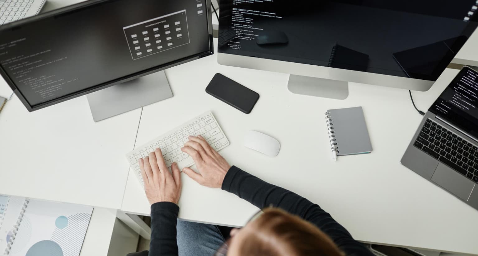 Person typing on a keyboard at a white desk with multiple monitors displaying code, a laptop, smartphone, notebook, and mouse nearby.