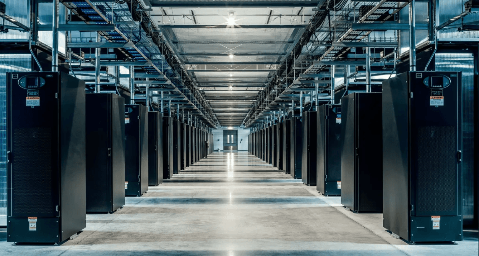 A long aisle in a server room with rows of black server racks under bright overhead lighting and visible cable trays above.