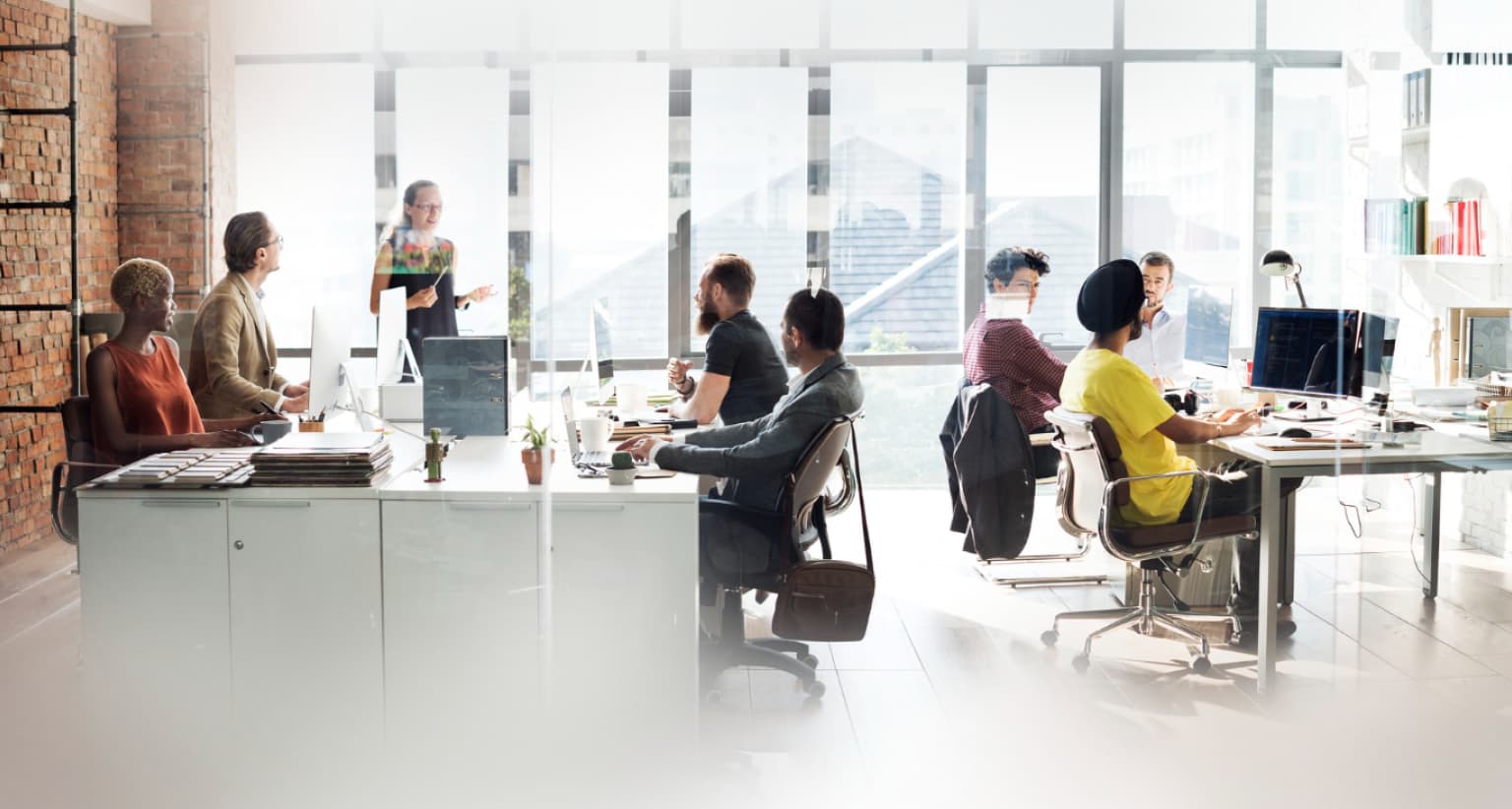 Open office scene with diverse employees working at desks, computers in use. One person stands, presenting to the team by large windows.