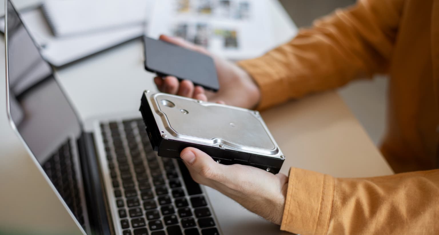 Person holding a hard drive and a smartphone near a laptop, wearing an orange sleeve.