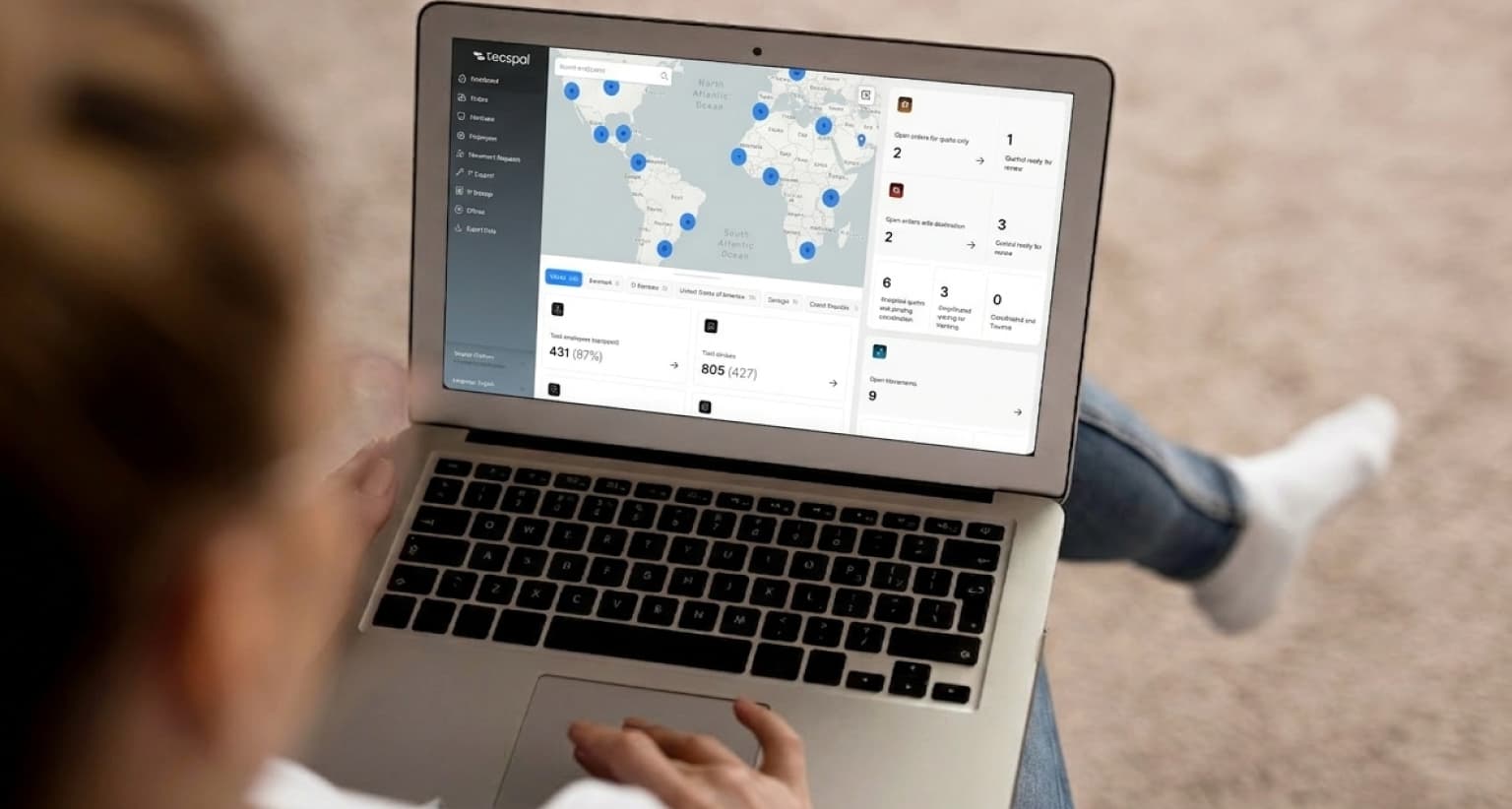Person using a laptop displaying a world map with location markers and data on the screen, sitting on a light-colored carpeted floor.