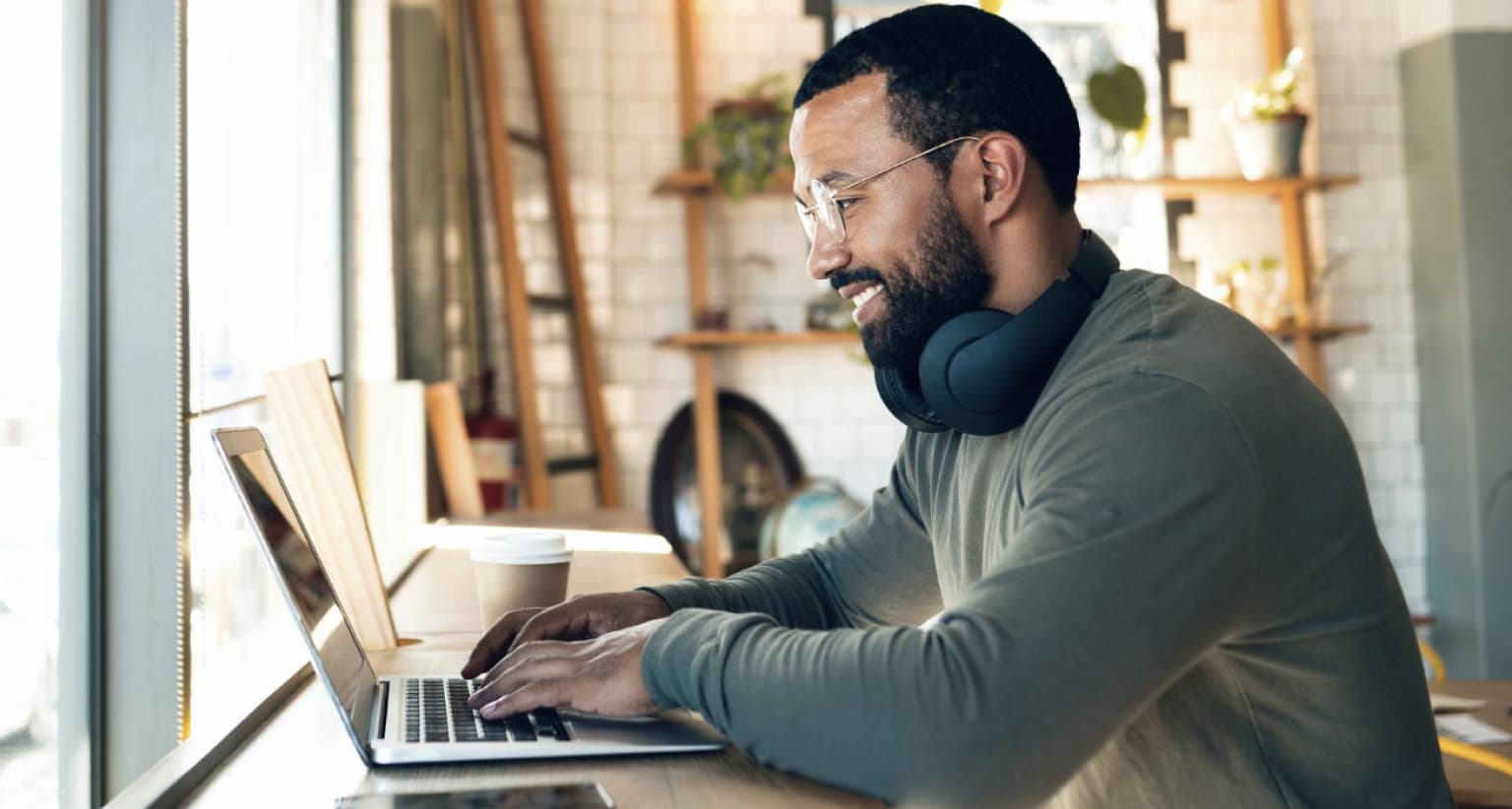 Man with headphones smiling while working on a laptop in a café, with a coffee cup nearby and shelves in the background.