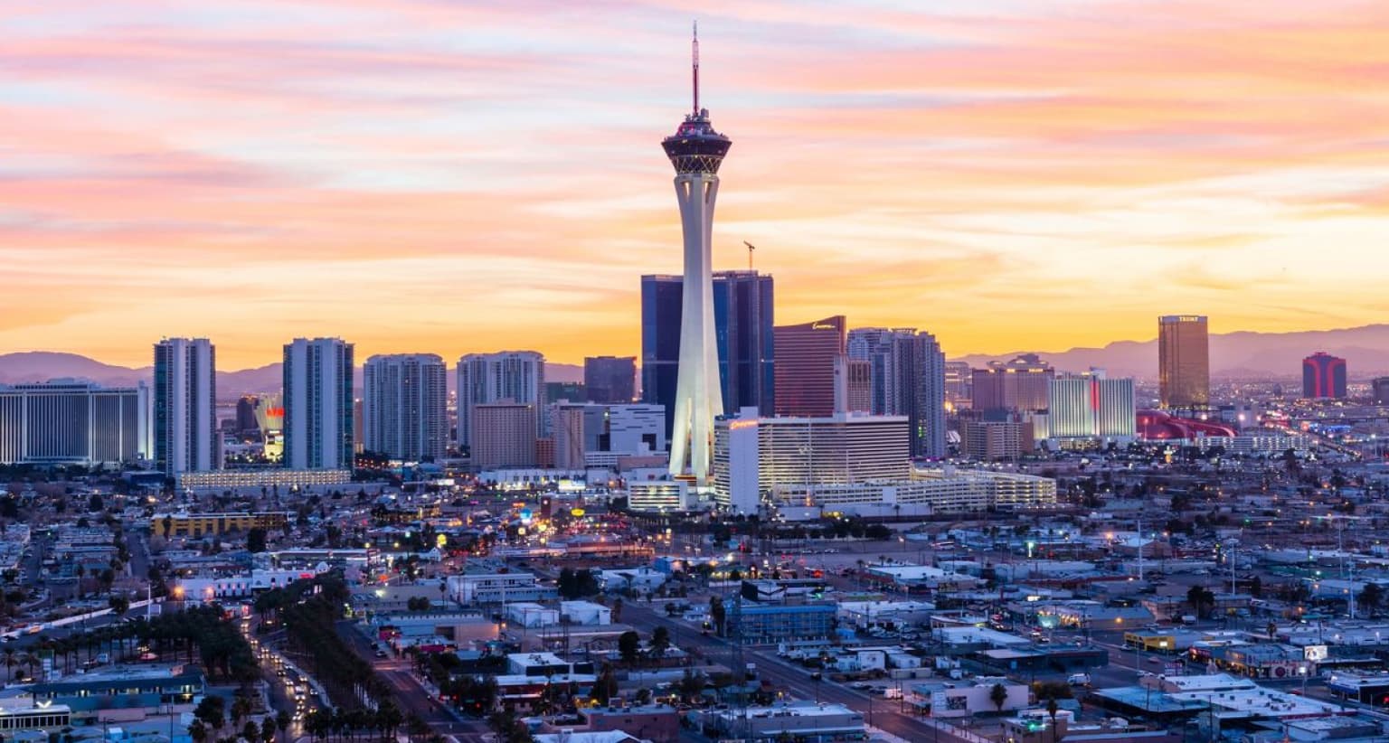 Las Vegas skyline at sunset, featuring the Stratosphere Tower surrounded by tall buildings and colorful sky in the background.