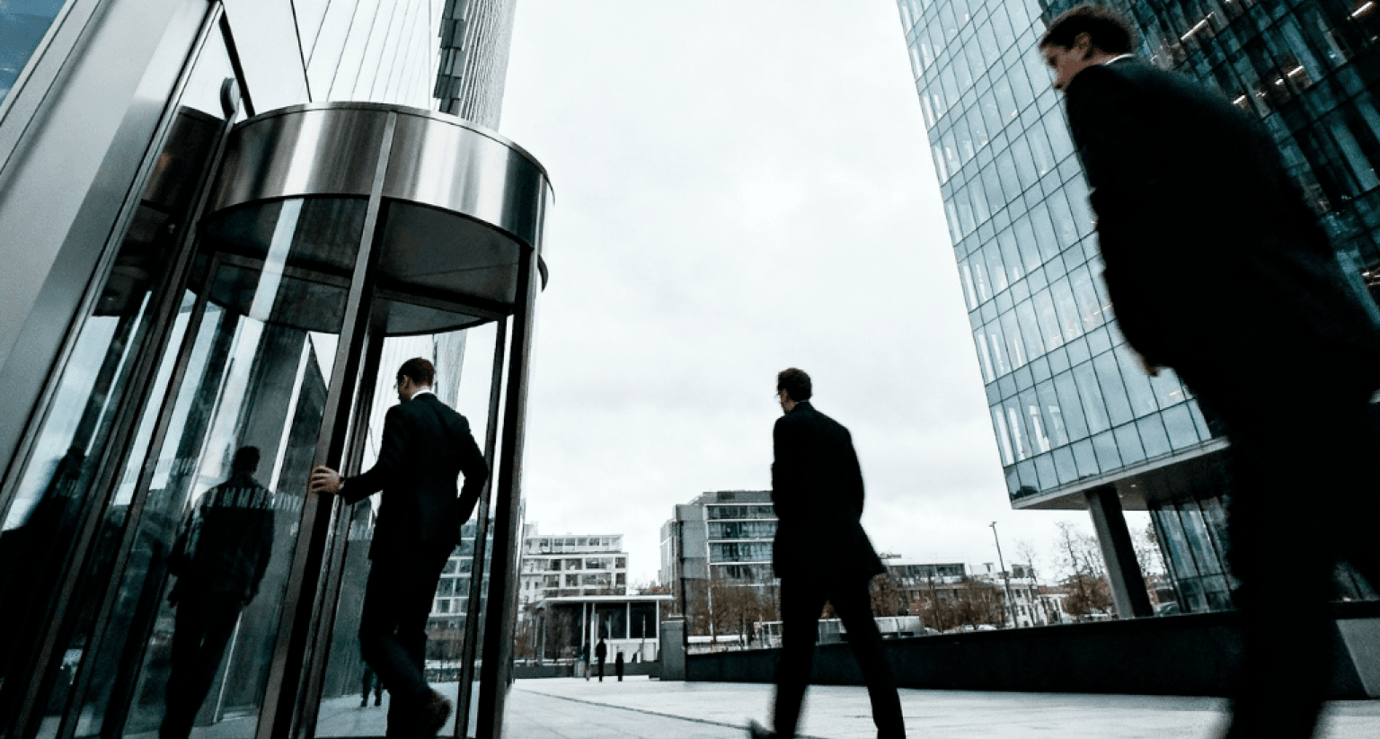 People in suits walking near modern glass buildings; one enters through a revolving door under a cloudy sky.