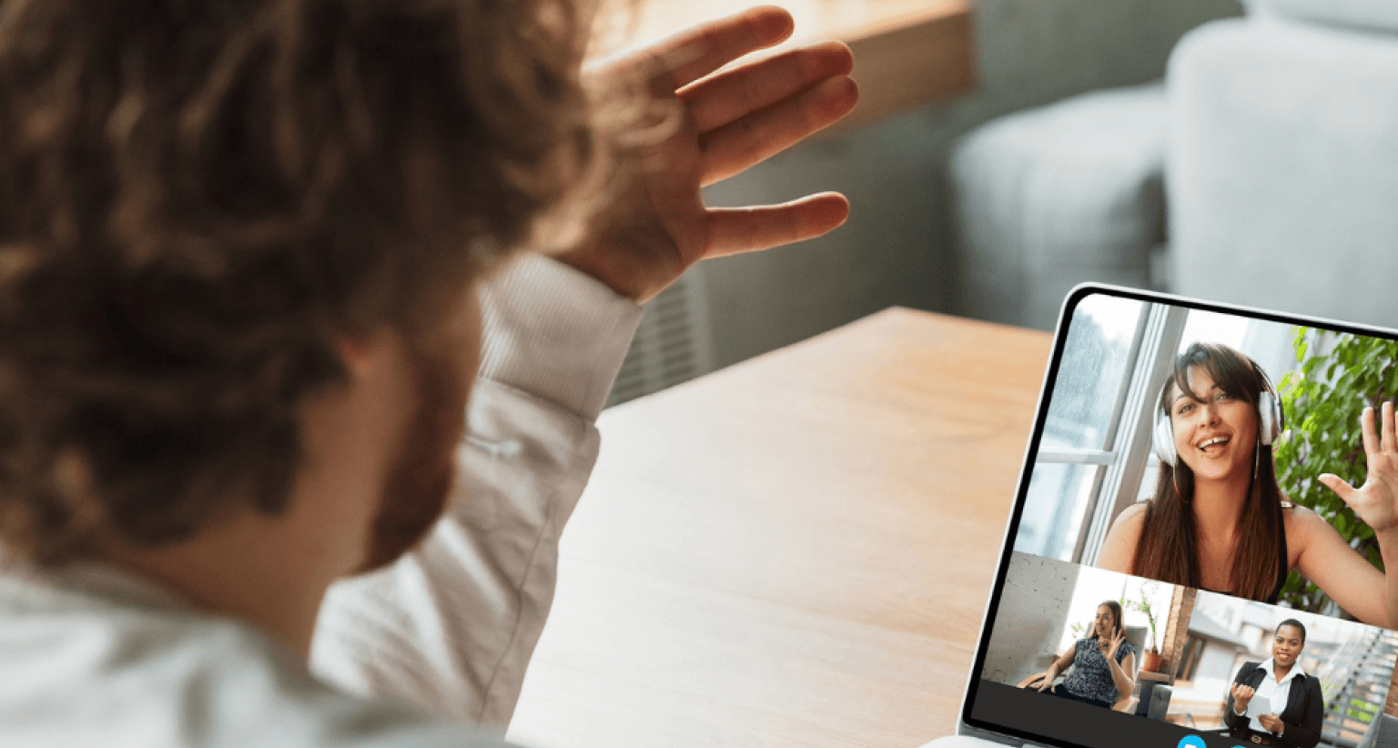 Person waves at a laptop screen during a video call with three people, one prominently visible smiling, in a cozy room setting.