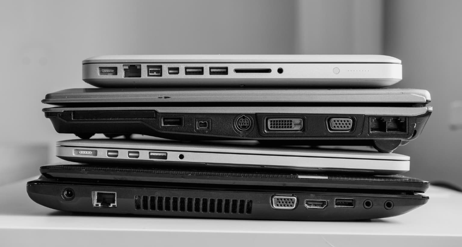 A black and white photo of four stacked laptops, showing various ports like USB, HDMI, and VGA on their sides.