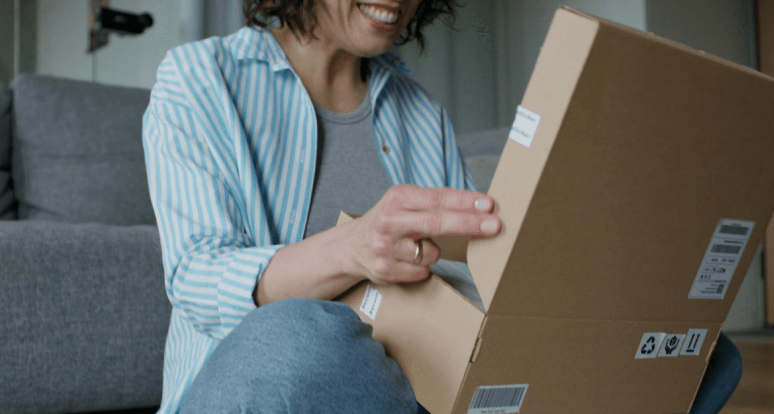 Smiling woman in striped shirt opens a cardboard box while seated on the floor near a couch.