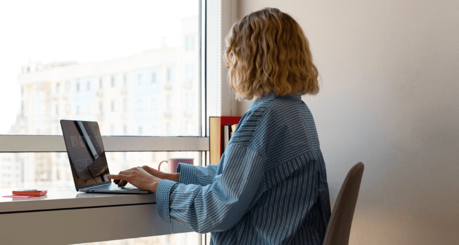 Woman with wavy hair using a laptop at a desk by a window, wearing a blue striped shirt, buildings visible outside.