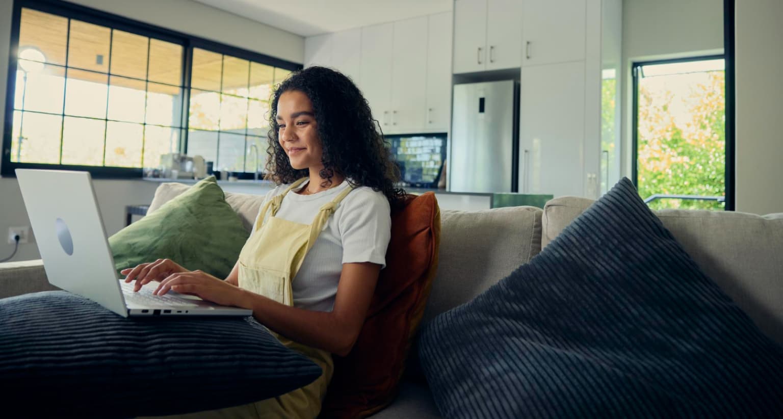 Woman sitting on a couch, using a laptop, in a bright living room with cushions and a view of the kitchen behind her.