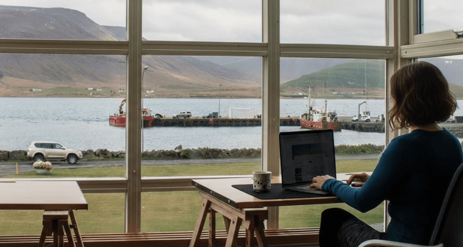 Woman working on a laptop by a large window, overlooking a scenic view of mountains, water, and a pier with vehicles and boats.