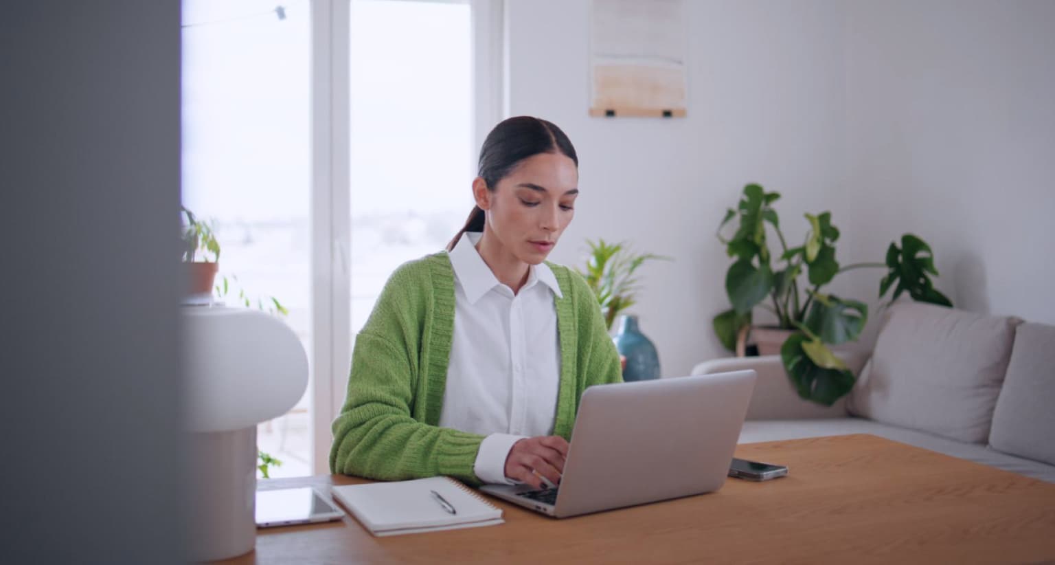 Woman in a green sweater works on a laptop at a wooden table, with plants and a sofa in the background, in a well-lit room.