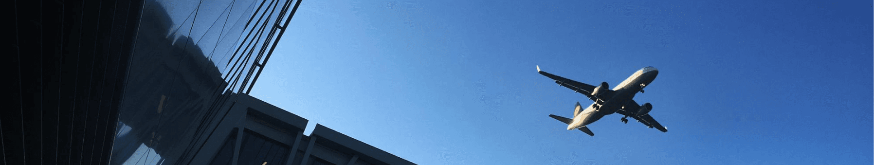Airplane flying above a modern building against a clear blue sky, with the building's glass facade reflecting light.
