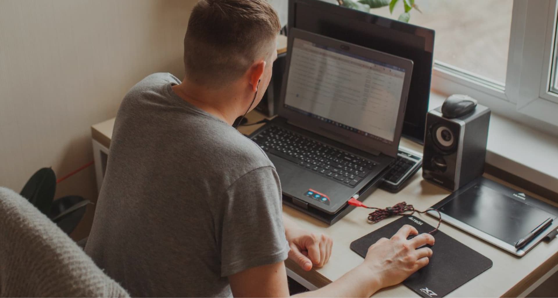 Person in a gray shirt sitting at a desk using a laptop, next to a phone, speakers, and notebook by a window.