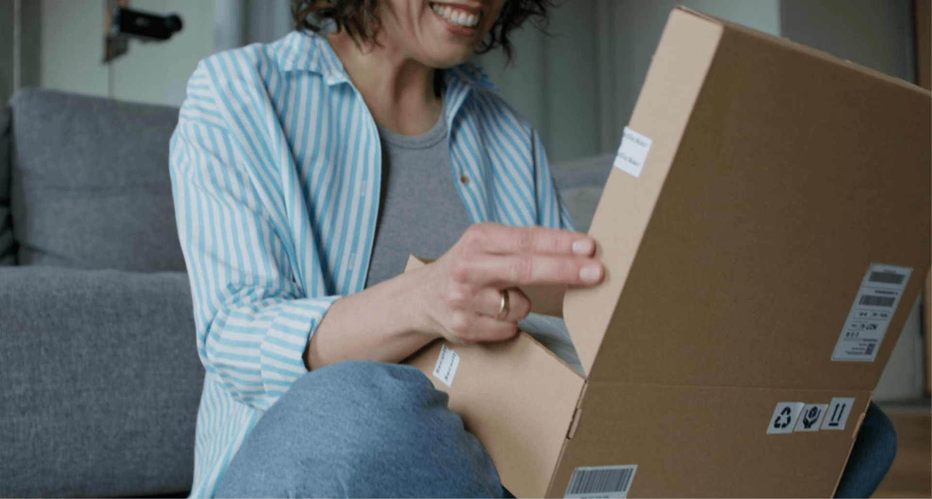 A person sitting on a couch, smiling while opening a cardboard package in a living room.