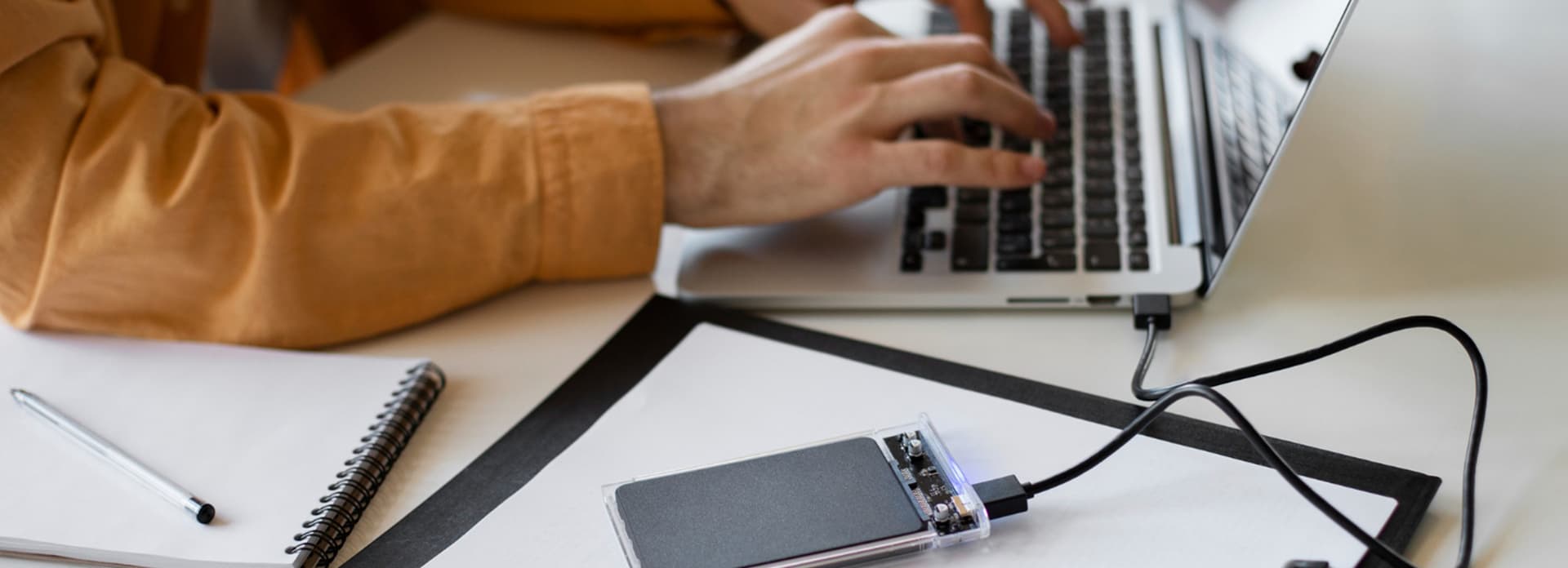 Person typing on a laptop with an external hard drive connected, notebook beside them on a desk.