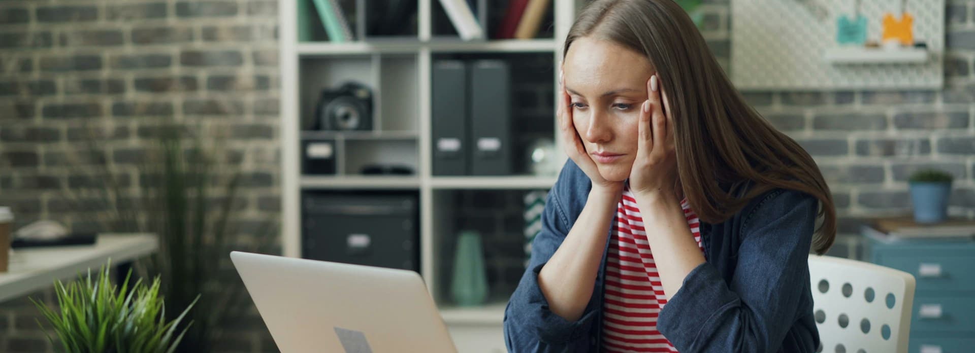 A woman with long hair, wearing a striped shirt and blue jacket, looks concerned while sitting at a desk, staring at a laptop in a home office.