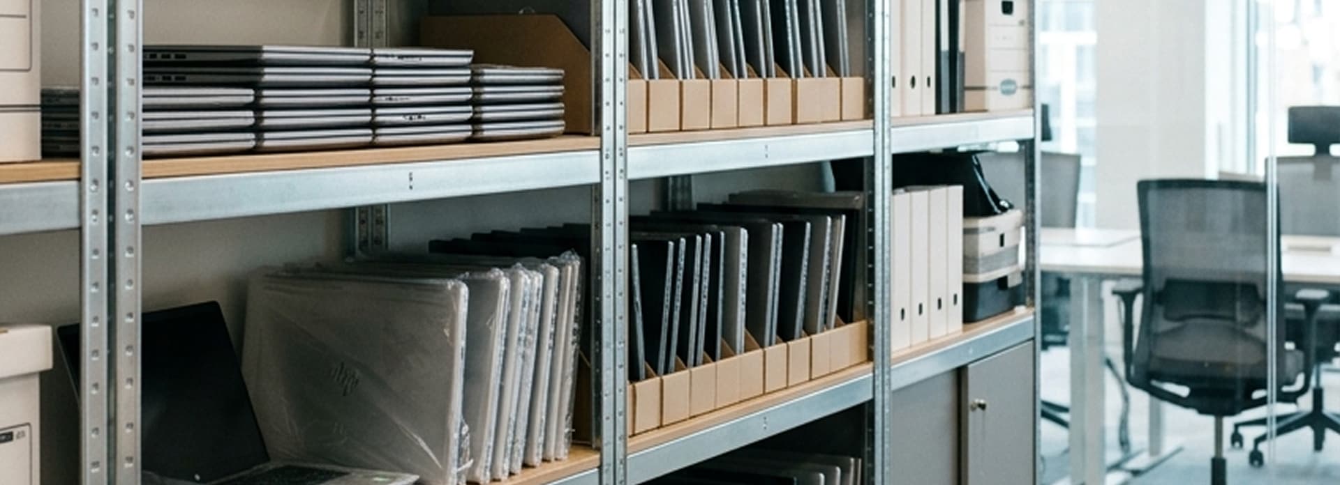 Office shelves with neatly stacked laptops, binders, and computer equipment in a modern workspace next to a glass-walled meeting room.