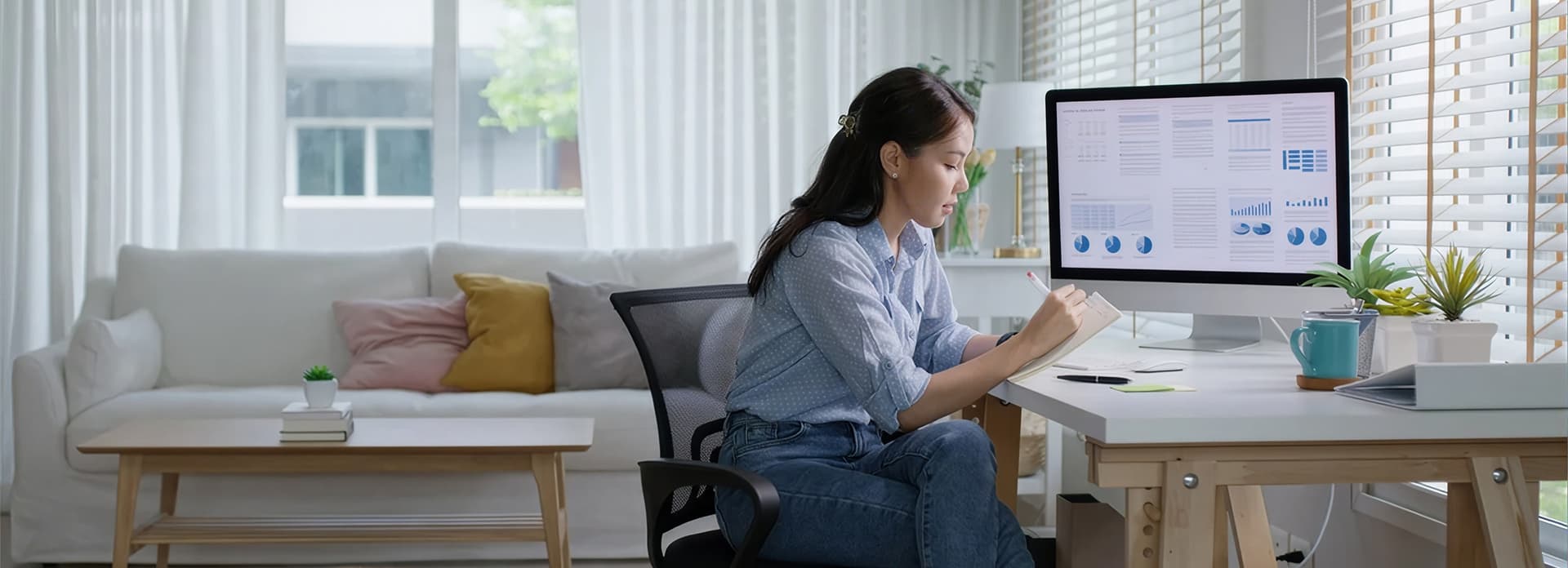 A woman in a blue shirt works at a desk with a large monitor displaying charts. A sofa with colorful cushions is in the background, near a window.
