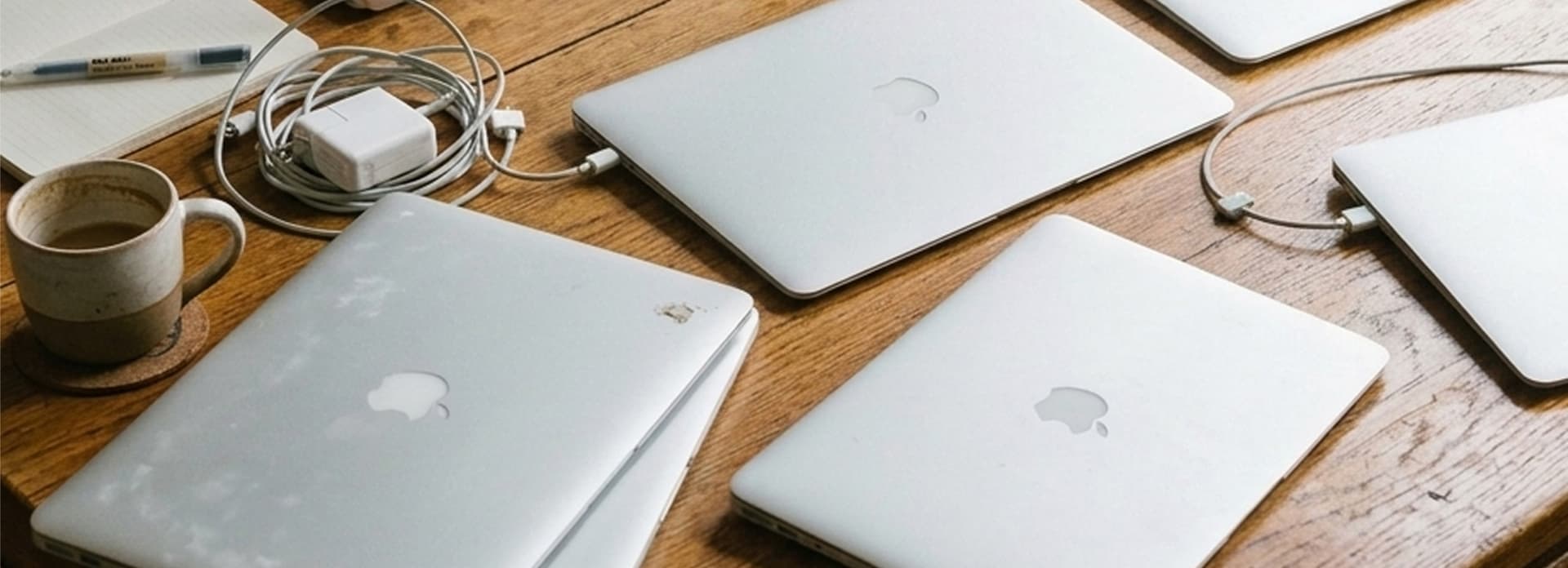 Several silver MacBooks on a wooden table, accompanied by a charger, notebook, pen, and coffee cup.