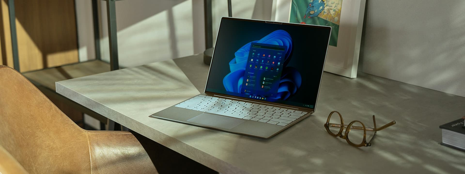 Laptop with a colorful screen on a sunlit desk next to a pair of glasses, with a chair and framed artwork in the background.