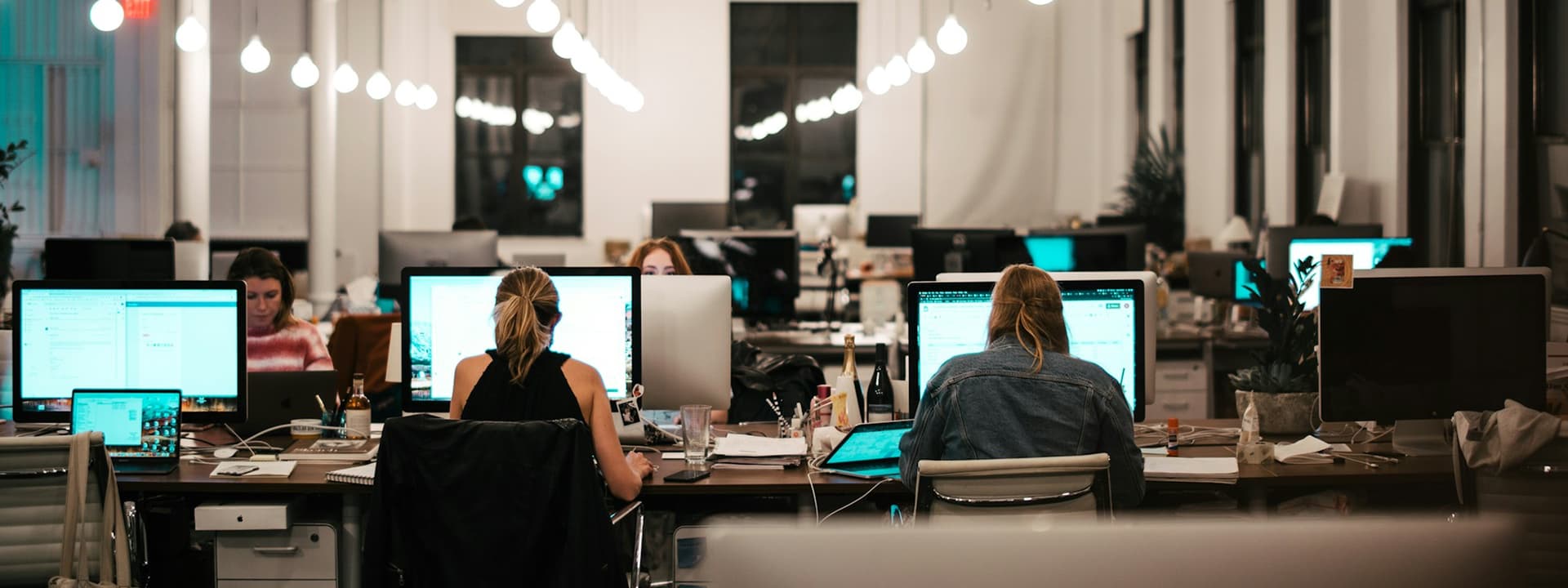 Open-plan office with people working at desks, illuminated by string lights. Multiple computers and chairs are visible, creating a busy workspace environment.