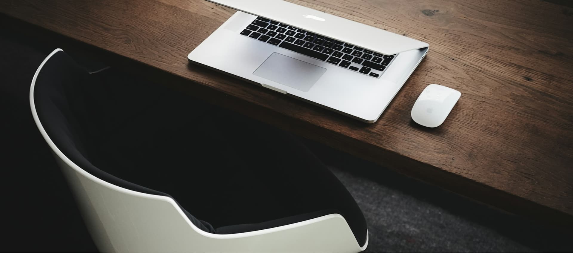 Open laptop and wireless mouse on a wooden desk, next to a curved white and black chair.