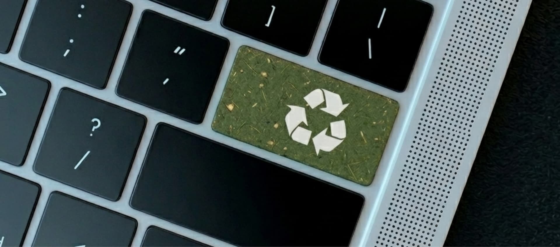 Laptop keyboard with a grass-textured key displaying a white recycling symbol, surrounded by regular black keys.