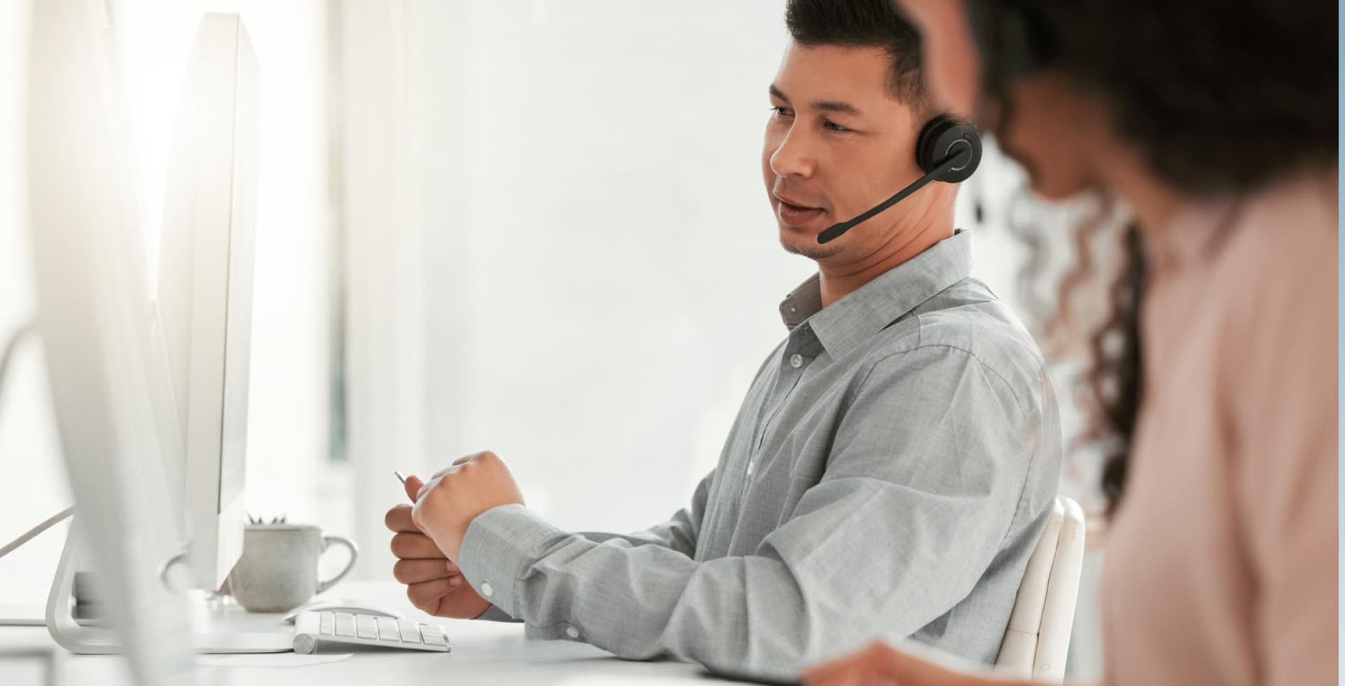 Man wearing a headset looks at a computer screen in a bright office, with another person blurred in the foreground.