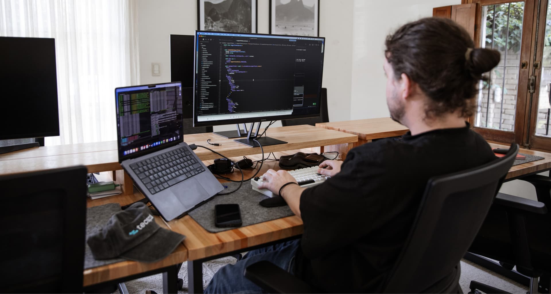 A person with long hair works on a dual-monitor setup, writing code on a computer, in a modern, well-lit room with wooden desks.