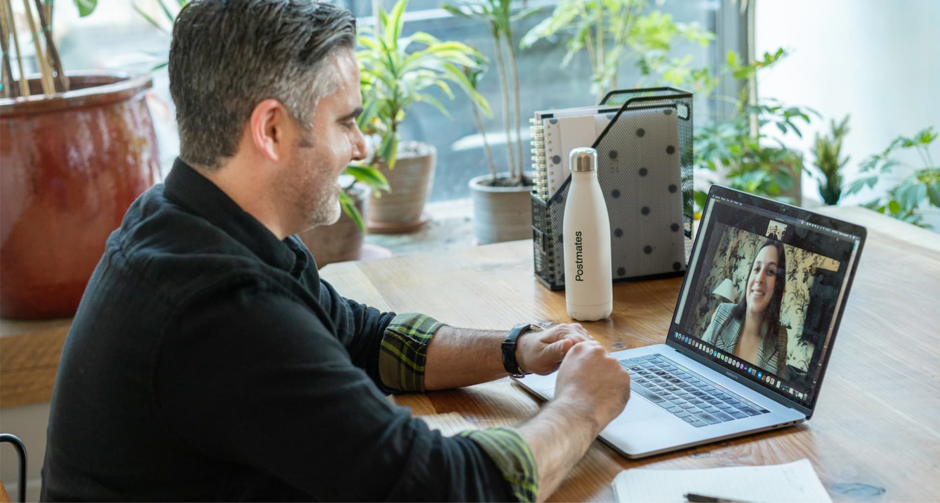 Man sitting at a wooden desk, having a video call on a laptop. A notebook and a reusable water bottle are on the desk, with plants in the background.