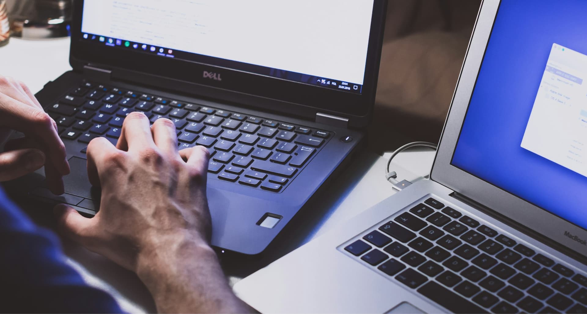 Two laptops on a table with someone's hands typing on one. The left laptop displays a code editor, and the right one shows an application window.