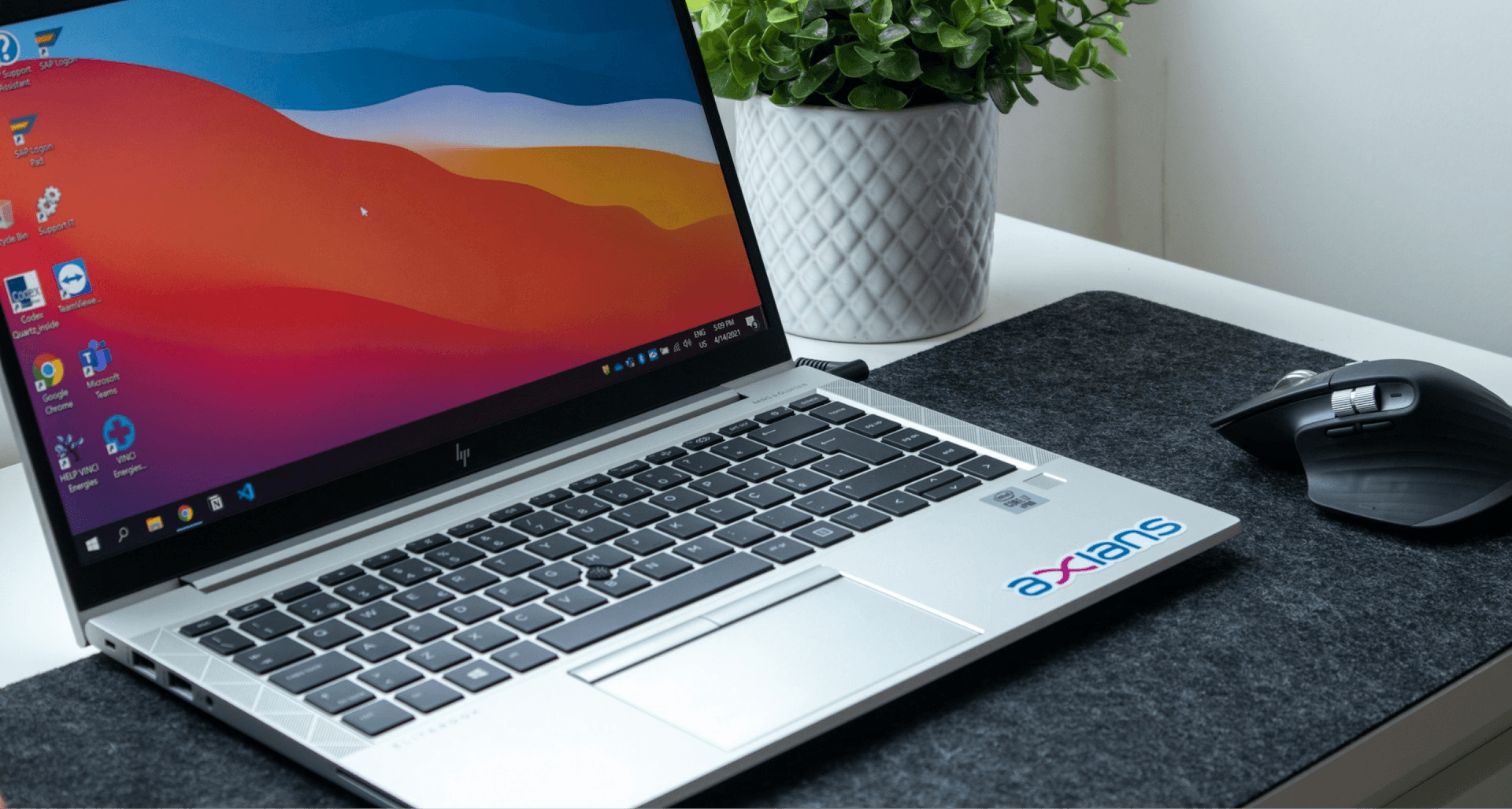 A laptop with a colorful screen on a desk, a plant in the background, and a wireless mouse on a black mat.