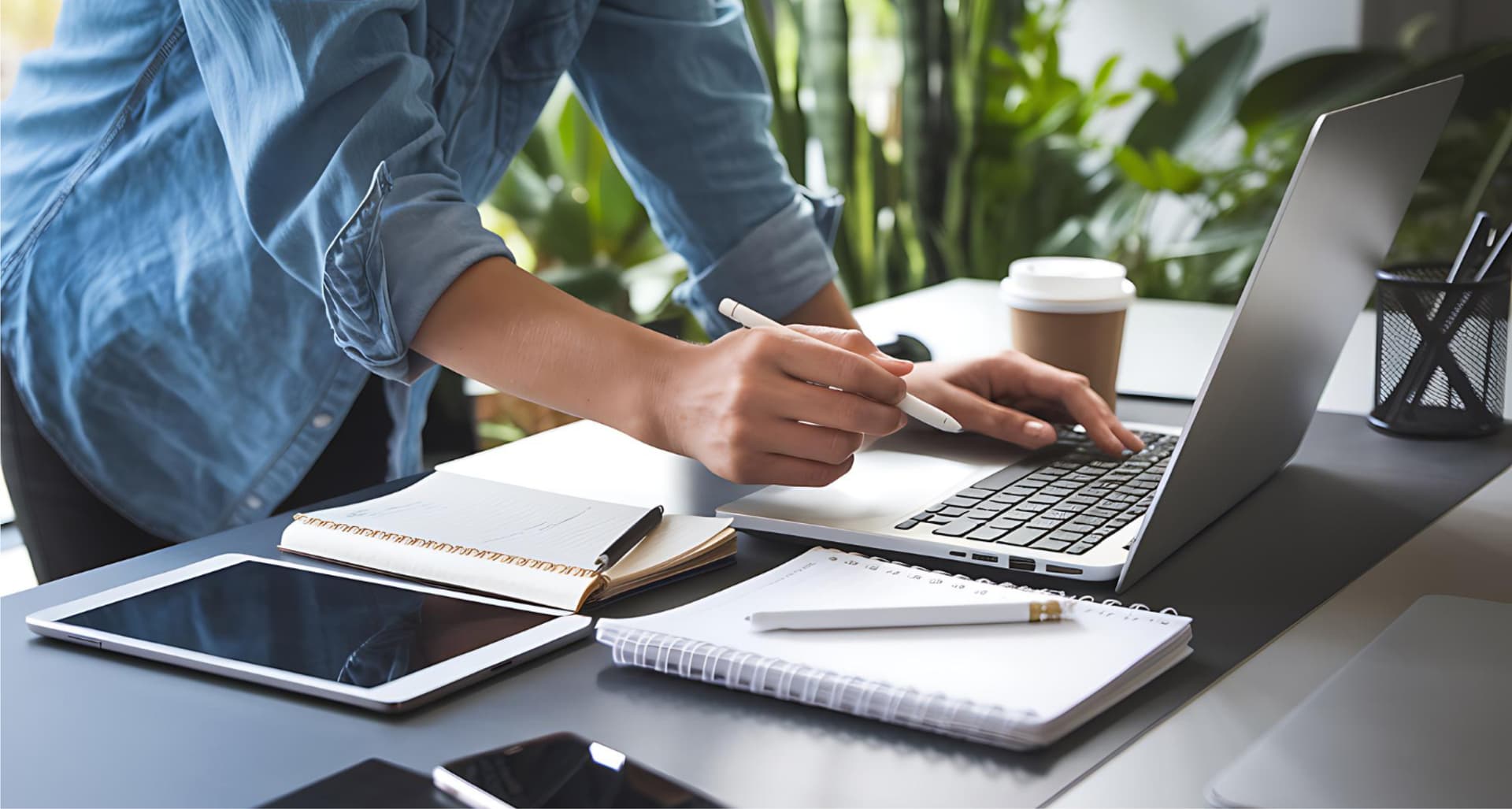 Person using a laptop at a desk with a tablet, phone, notebooks, and a coffee cup, surrounded by greenery.