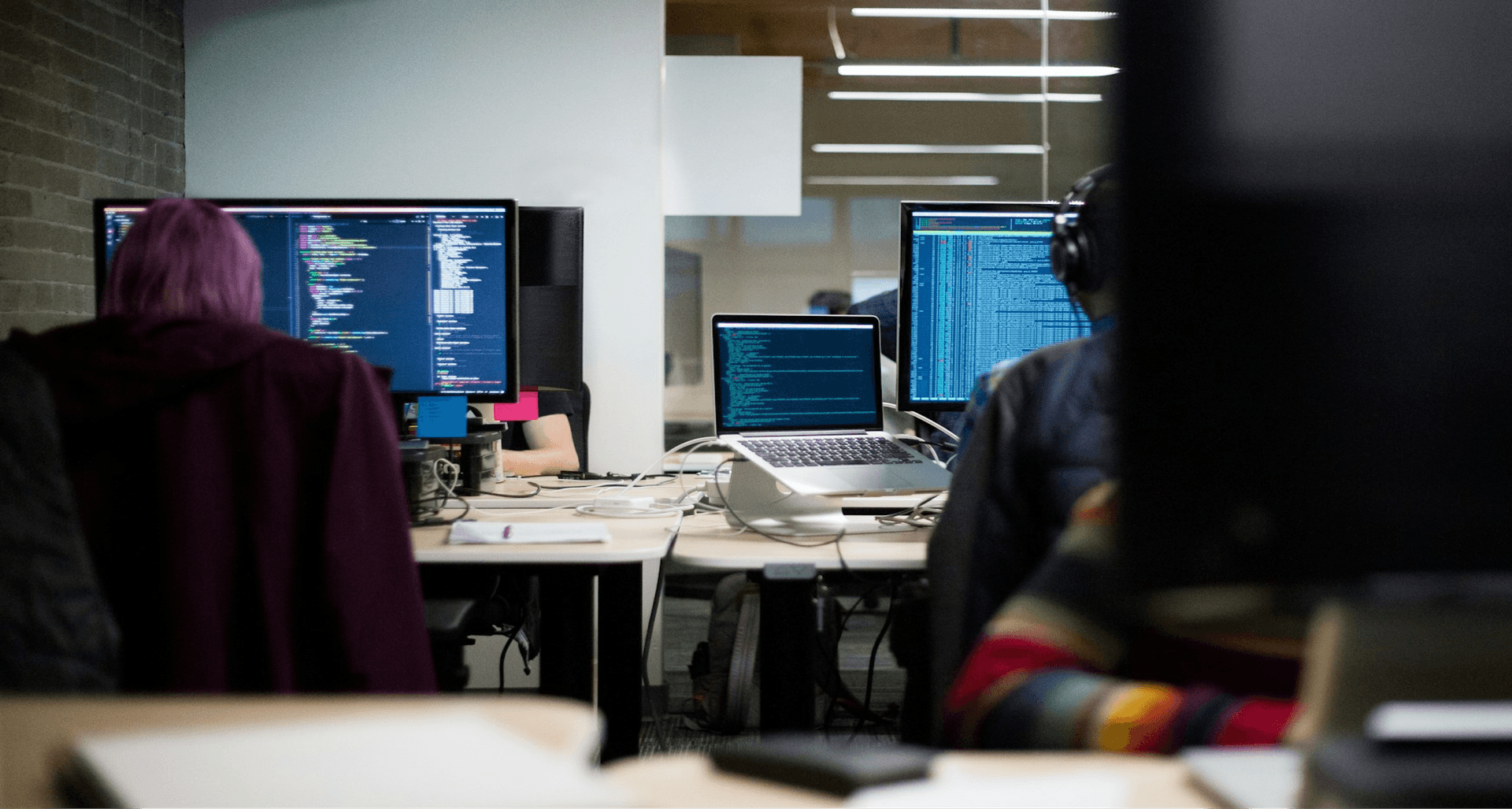 People working at desks in a dimly lit office, focused on coding on multiple computer screens.