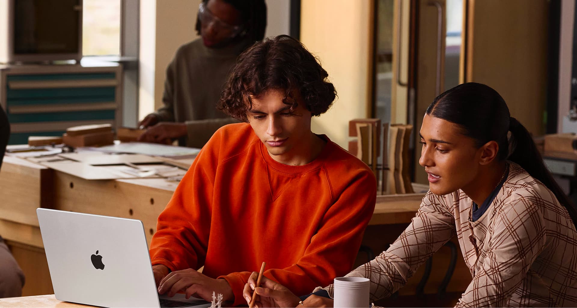 Two people collaborate at a table, looking at a laptop. One wears an orange sweater, the other a checked shirt. A coffee cup and tools are nearby.