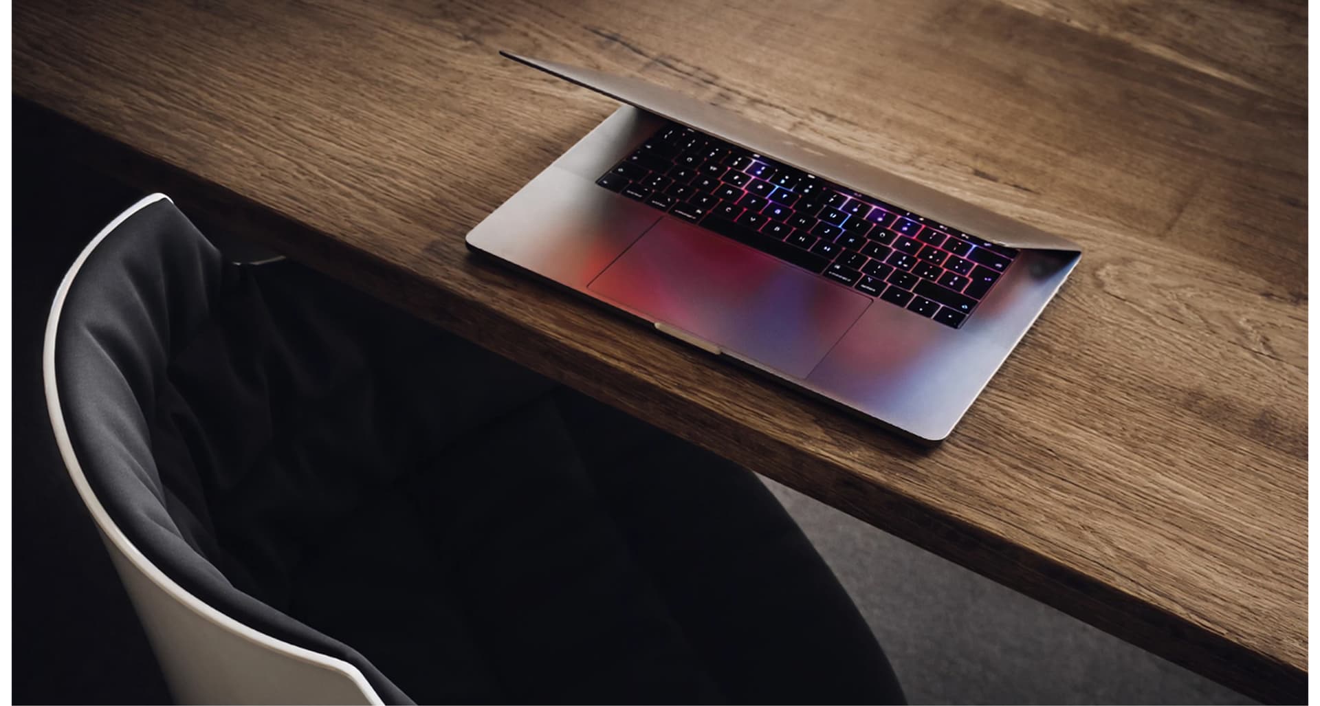 A partially closed laptop with a glowing keyboard on a wooden desk, next to a modern white and black chair.