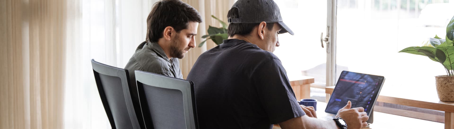 Two people sitting at a table by a window, focused on a laptop. One is wearing a cap; plants are visible in the background.