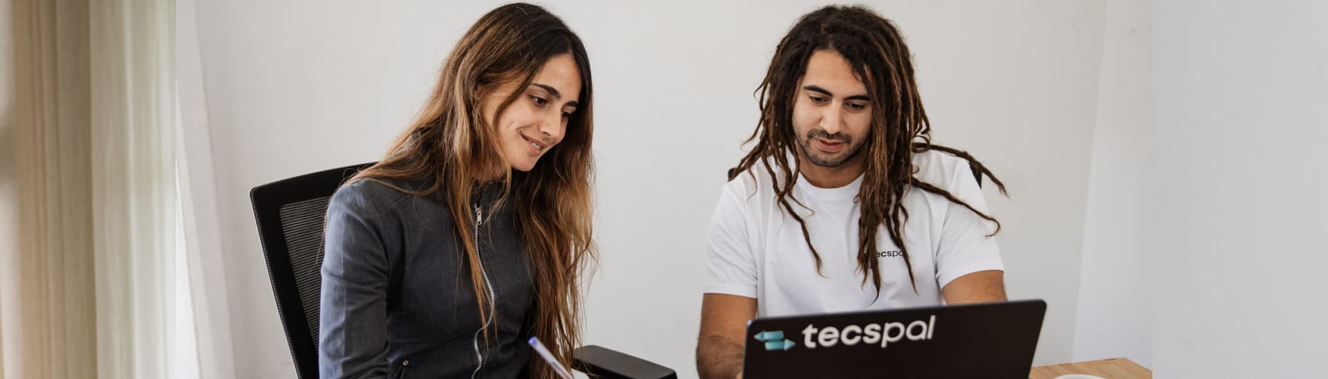 A man and a woman sitting at a desk, looking at a laptop. The woman is taking notes, and the man is typing.