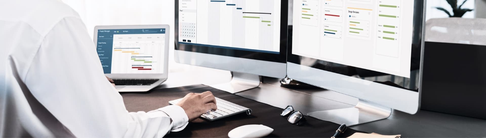 Person working at a desk with multiple computer screens displaying scheduling and planning software, using a keyboard and mouse.