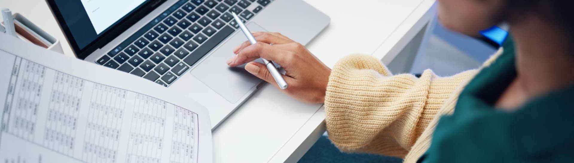 Person in a sweater working on a laptop at a desk, holding a pen and reviewing a printed spreadsheet.