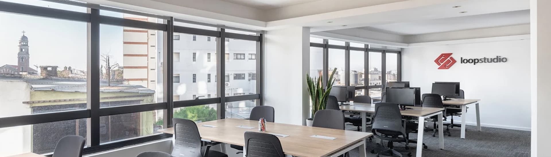 Modern office with large windows, bright natural light, and desks with chairs. "Loopstudio" logo on the wall. Urban view in the background.