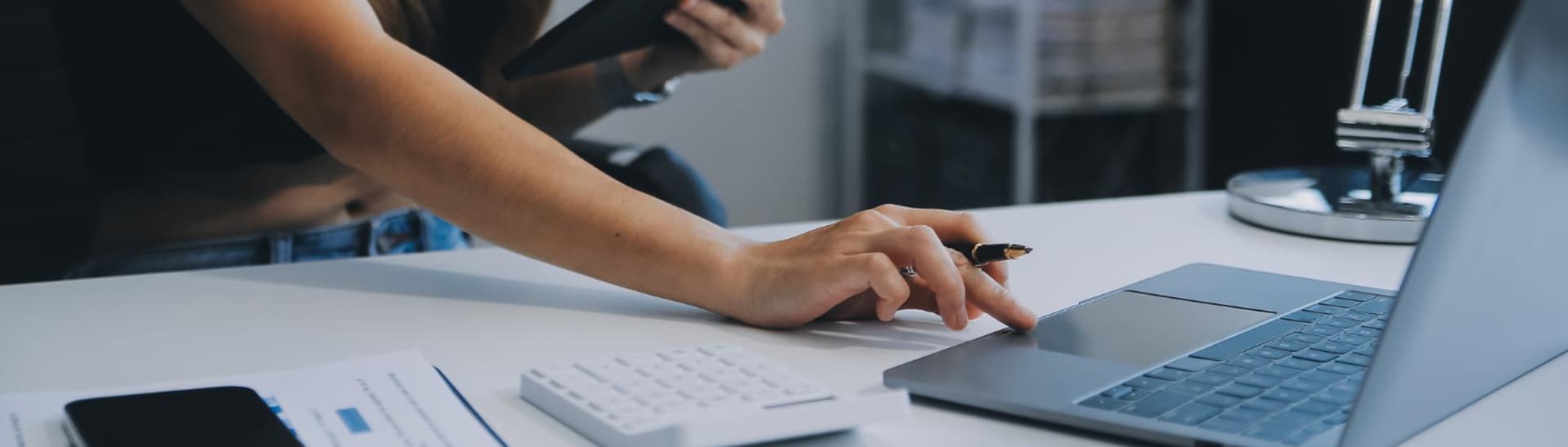 Person working on a laptop at a desk, with a calculator and documents nearby, another person holding a tablet in the background.