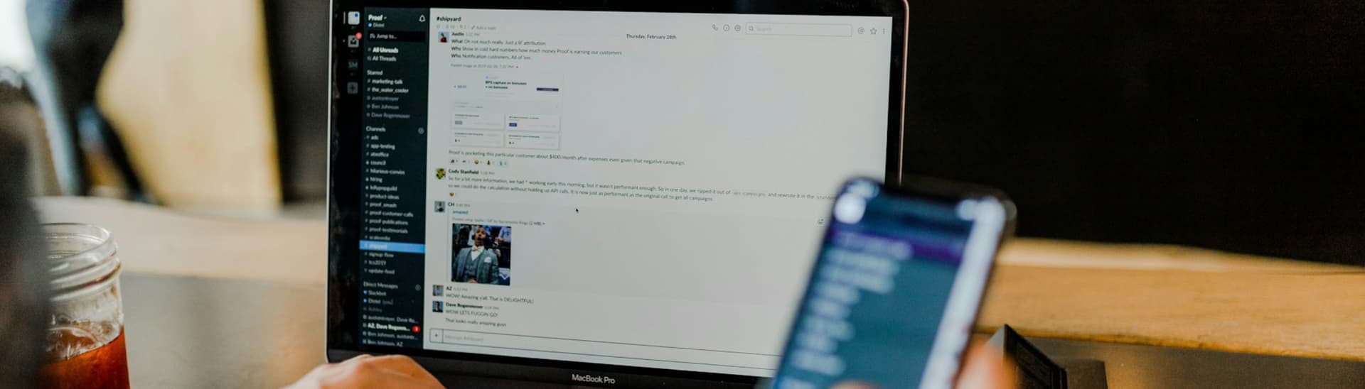 Person using a smartphone and laptop with a messaging app open, indicating multitasking. A beverage is on the table beside the laptop.
