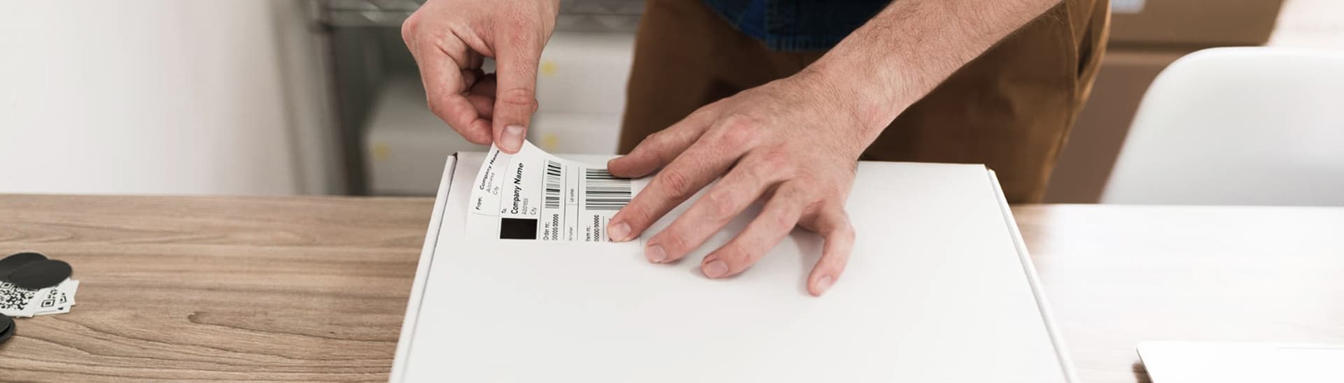 Person applying a shipping label to a white box on a wooden table.