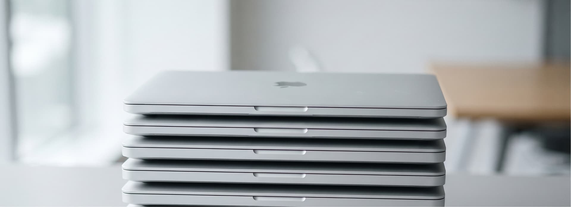 A stack of closed silver laptops on a table in a bright, modern office setting.