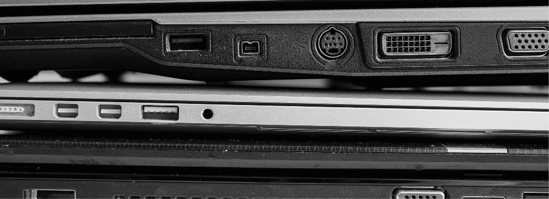 Stack of assorted laptops focusing on side ports, including USB, HDMI, and VGA connectors. Black and white image emphasizes hardware detail.